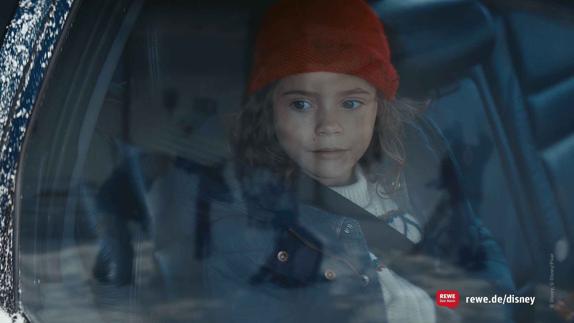 Still from the REWE Disney commercial – a girl in a red knitted hat looking out through a car window in winter 