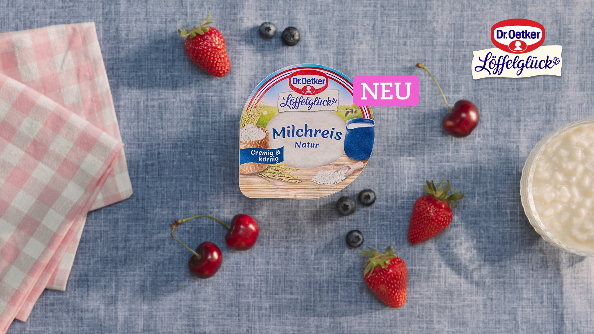 Still from the Dr. Oetker Löffelglück commercial – a Milchreis Natur pot on blue linen surrounded by strawberries, cherries and blueberries 