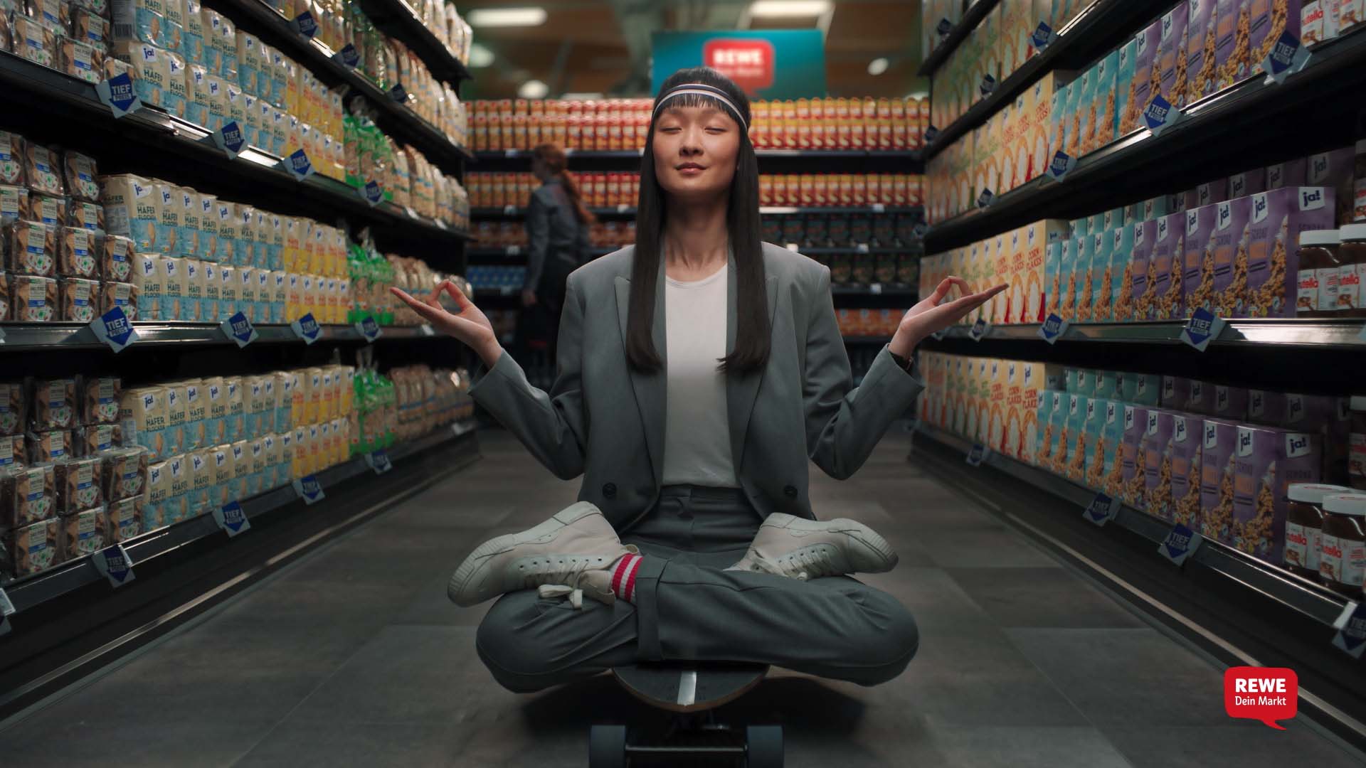 Still from the REWE Tiefpreis commercial – woman in a grey suit meditating cross-legged on a longboard in the cereal aisle 