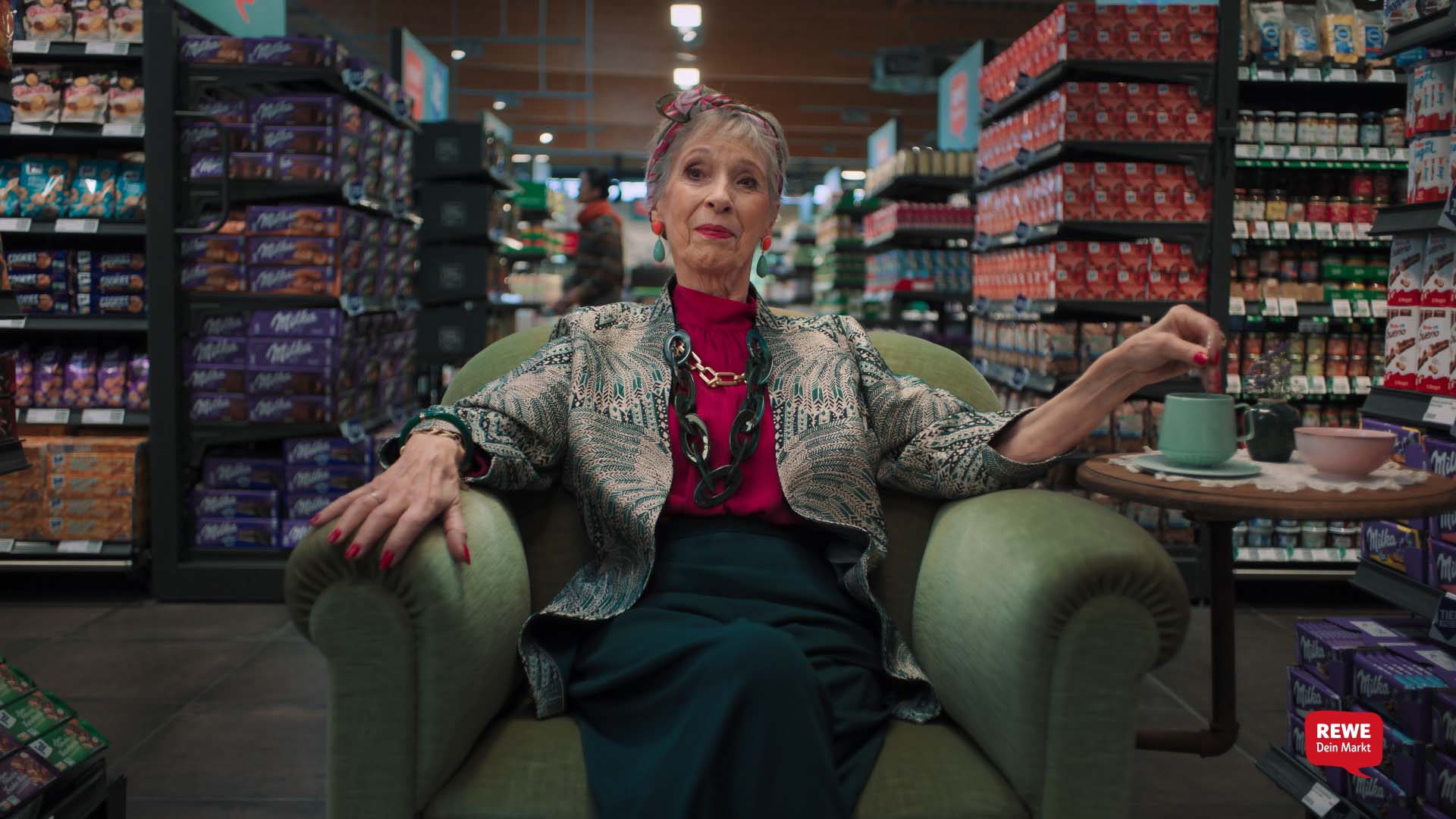 Still from the REWE Tiefpreis commercial – older woman sitting in an armchair with a cup of tea in the confectionery aisle 