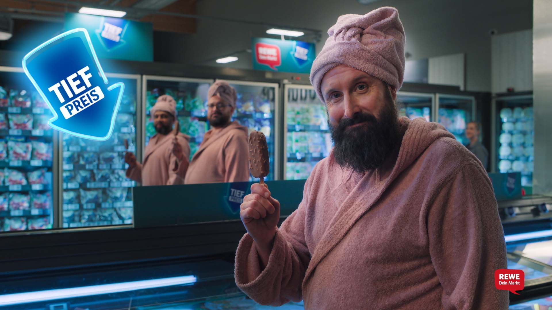 Still from the REWE Tiefpreis commercial – man in a pink bathrobe and towel turban holding a chocolate ice cream bar in the freezer aisle 