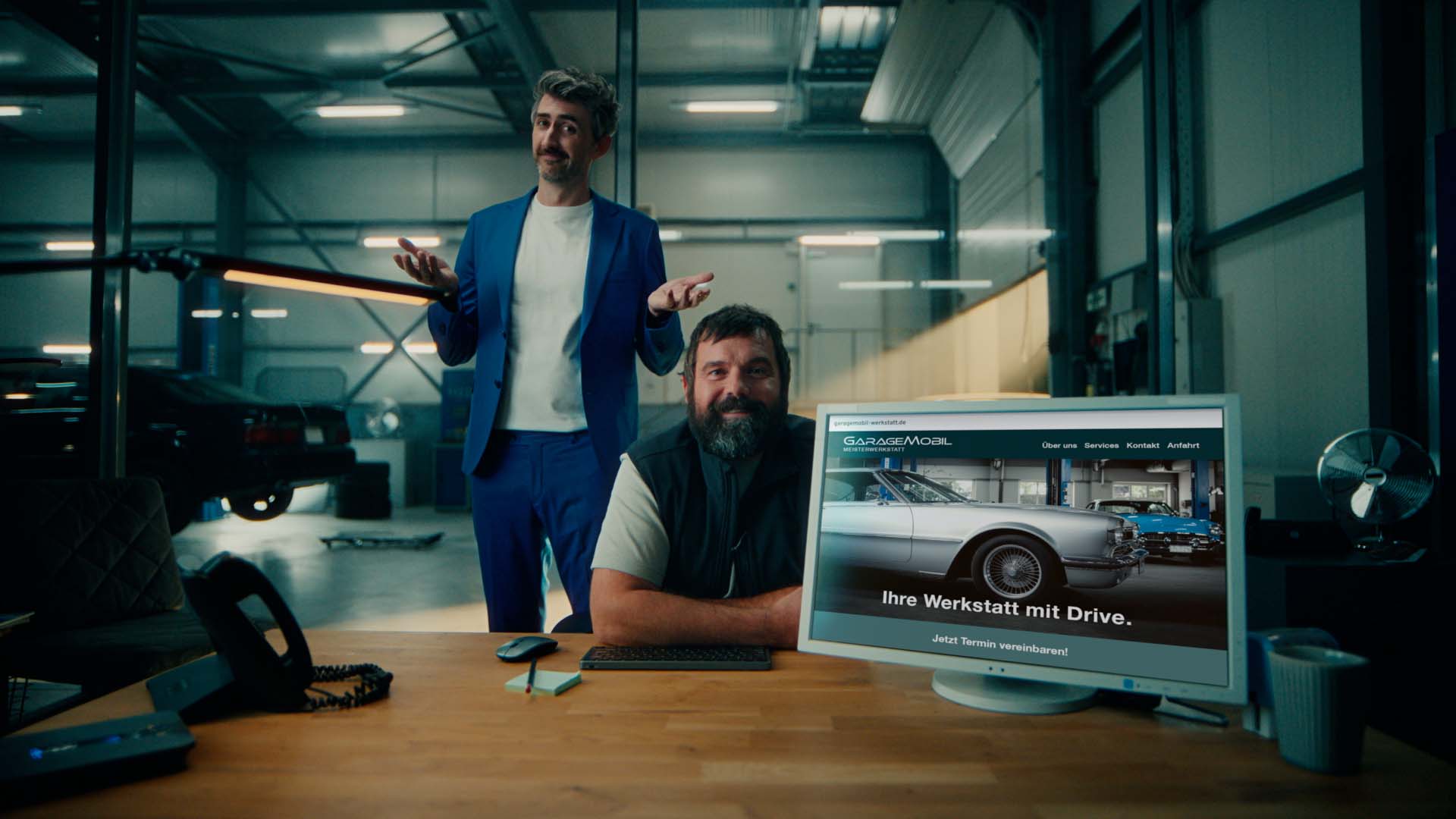 Still from the IONOS commercial – mechanic at a workshop desk beside a monitor showing a garage website 