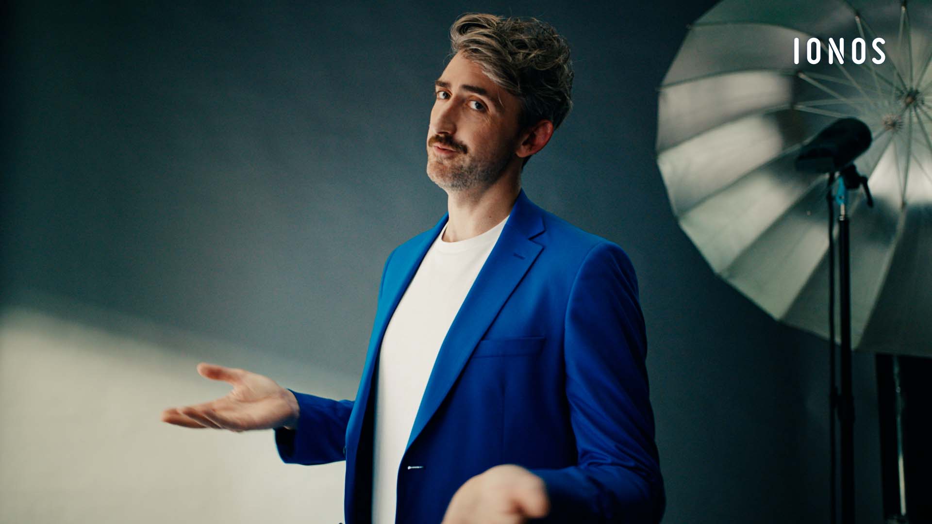 Still from the IONOS brand commercial – man in a blue suit gesturing beside a lighting umbrella in a photo studio 